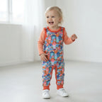 A toddler stands in a bright, minimalist white room wearing a long-sleeve orange shirt and colorful patterned baby dungarees with white sneakers.