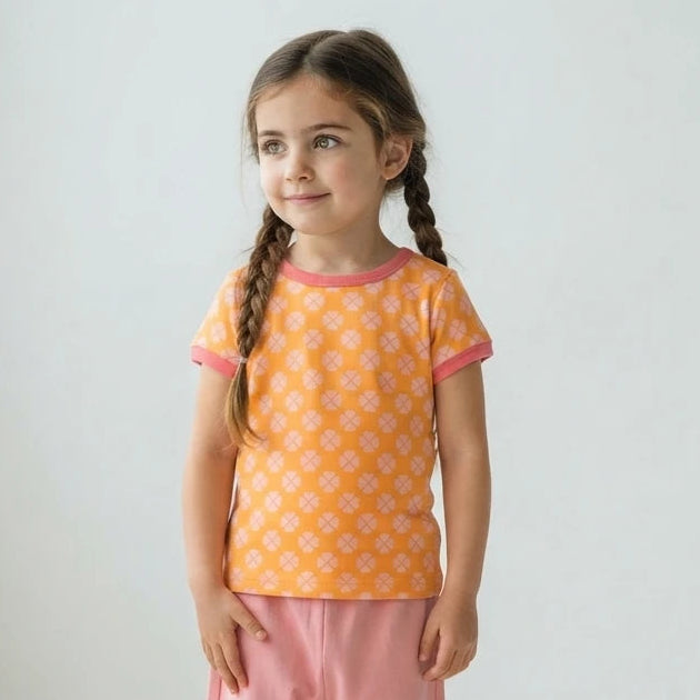 A young girl with braided hair stands in a bright white room wearing a short-sleeve orange t-shirt with a white geometric pattern and pink trim, paired with pink pants and white sneakers.