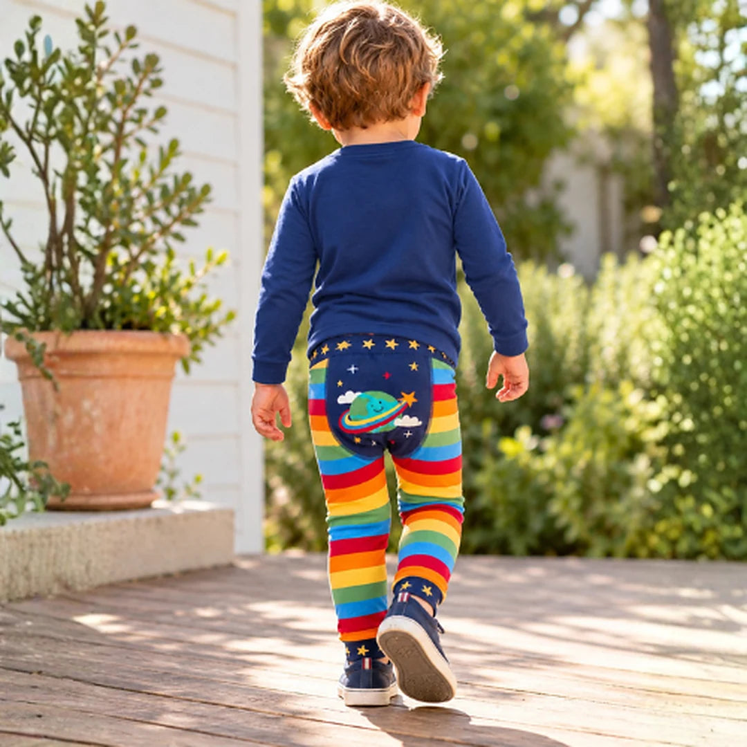 Toddler walking away in the Frugi Little Knitted Leggings in rainbow stripe with the smiling planet on the back