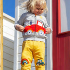 Child wearing a hoodie with a car design and yellow pants with vehicle patches, standing in front of a colorful background.