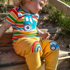 Child wearing a colorful striped shirt and yellow pants with car designs, sitting on a wooden bench.