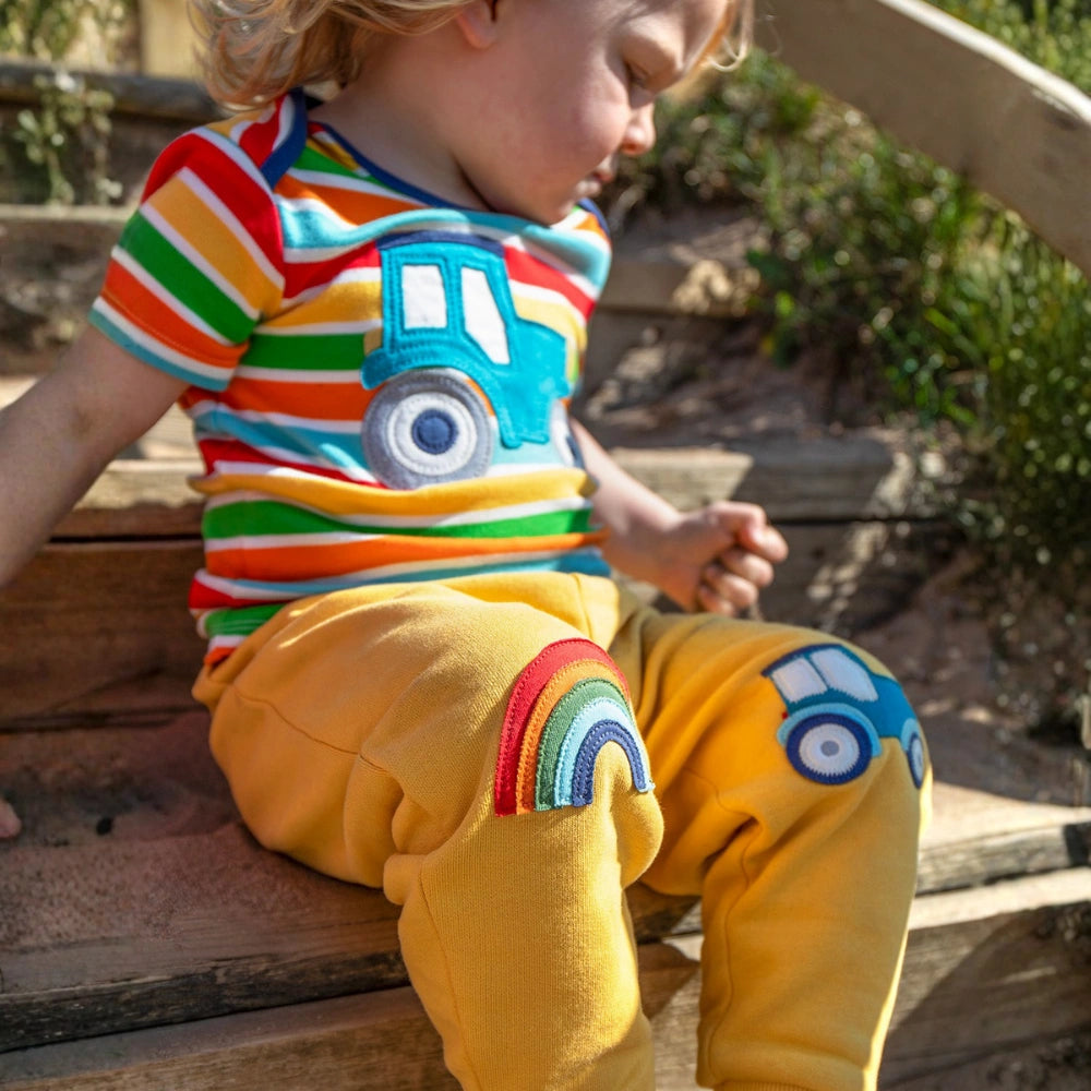 Child wearing a colorful striped shirt and yellow pants with car designs, sitting on a wooden bench.