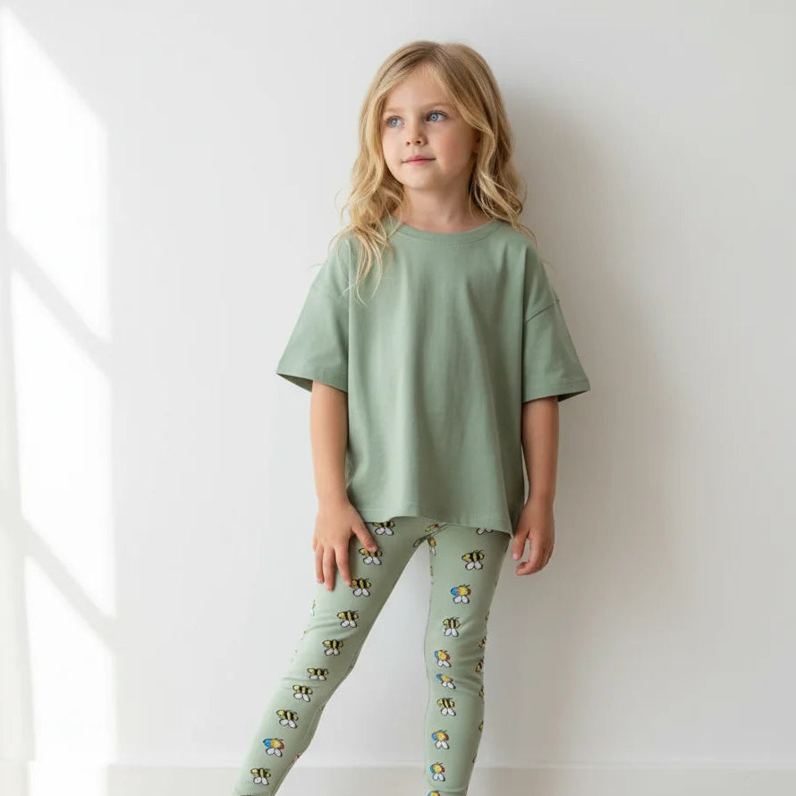 Young girl wearing a green outfit with patterned leggings against a white wall.