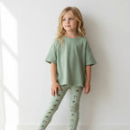 Young girl wearing a green outfit with patterned leggings against a white wall.
