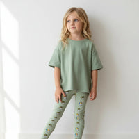 Young girl wearing a green outfit with patterned leggings against a white wall.