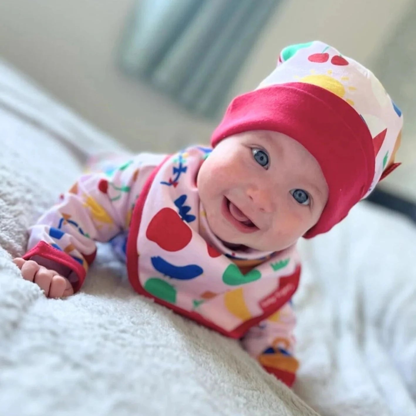 Baby wearing a colorful outfit and red hat on a light-colored blanket