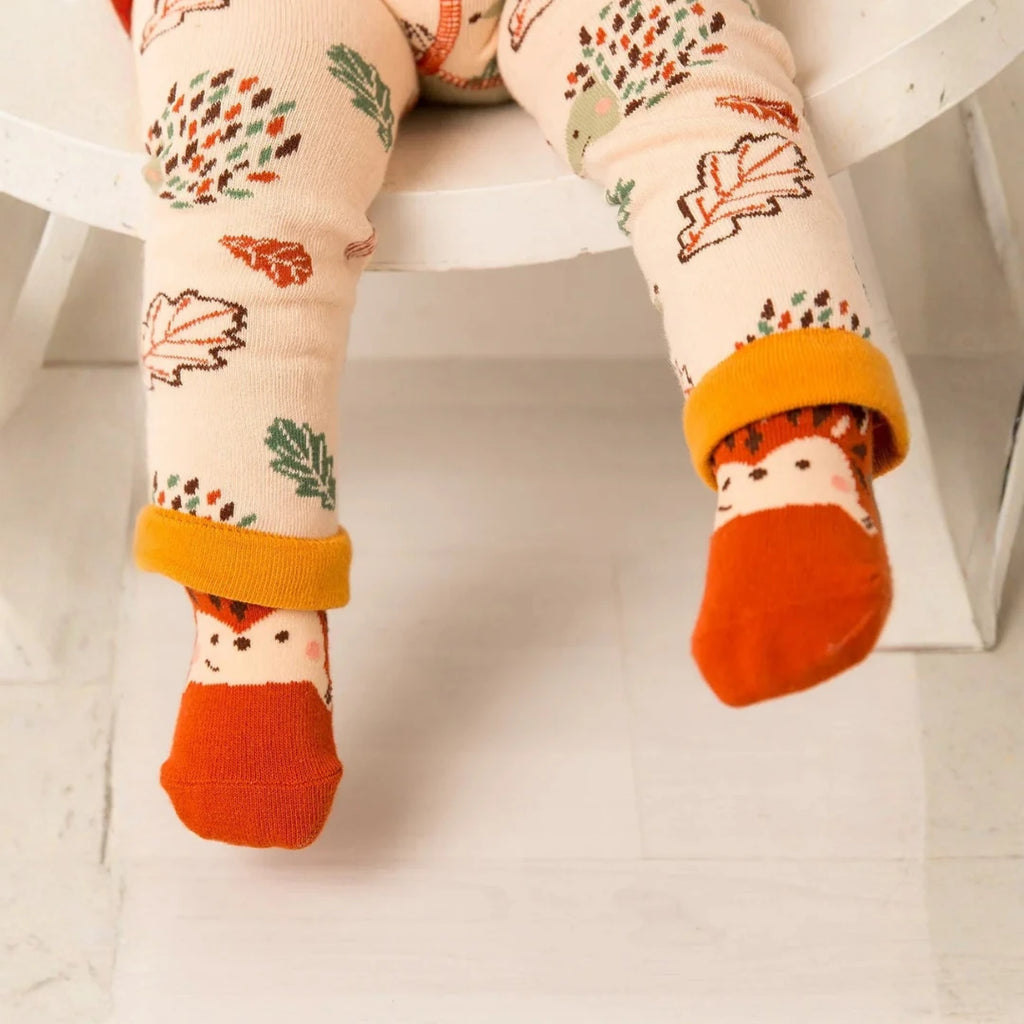 A close-up of a child's legs wearing cream-colored tights with an autumn leaf pattern and bright orange socks featuring hedgehog faces, sitting on a white wooden chair.