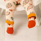 A close-up of a child's legs wearing cream-colored tights with an autumn leaf pattern and bright orange socks featuring hedgehog faces, sitting on a white wooden chair.