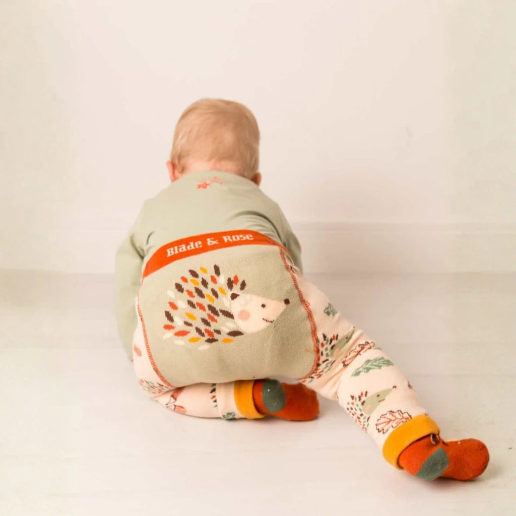 A baby sitting on the floor, seen from behind, wearing a sage green top and patterned leggings with orange socks. The back of the top features a large hedgehog graphic with the brand name 'Blade & Rose' printed on an orange band.