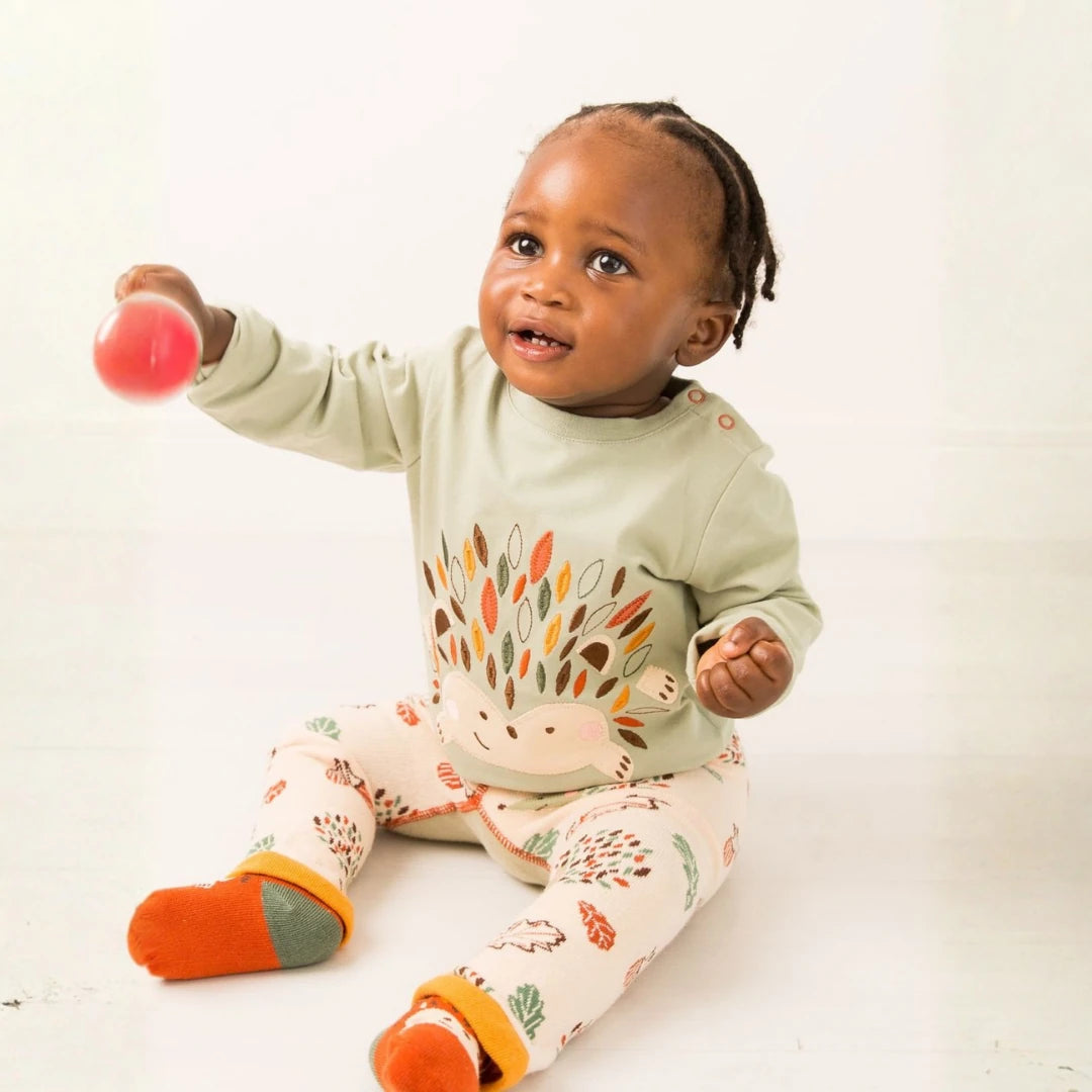 A baby sitting on a white floor wearing a sage green long-sleeve top with a hedgehog design and cream leggings with colorful leaf prints. The baby is holding a pink ball.