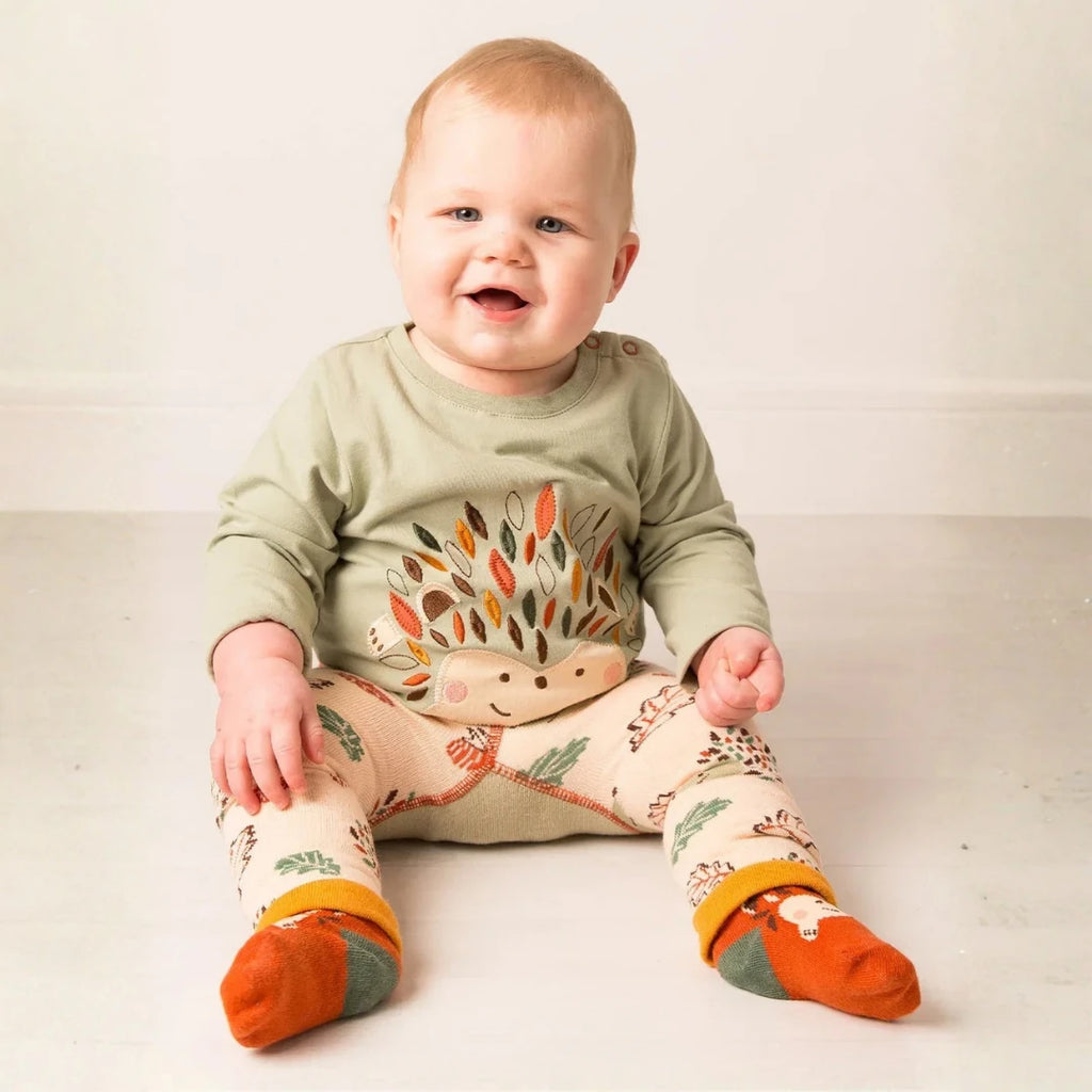 A baby sitting on a light floor wearing a sage green long-sleeve top with a hedgehog graphic, cream patterned pants, and orange socks.