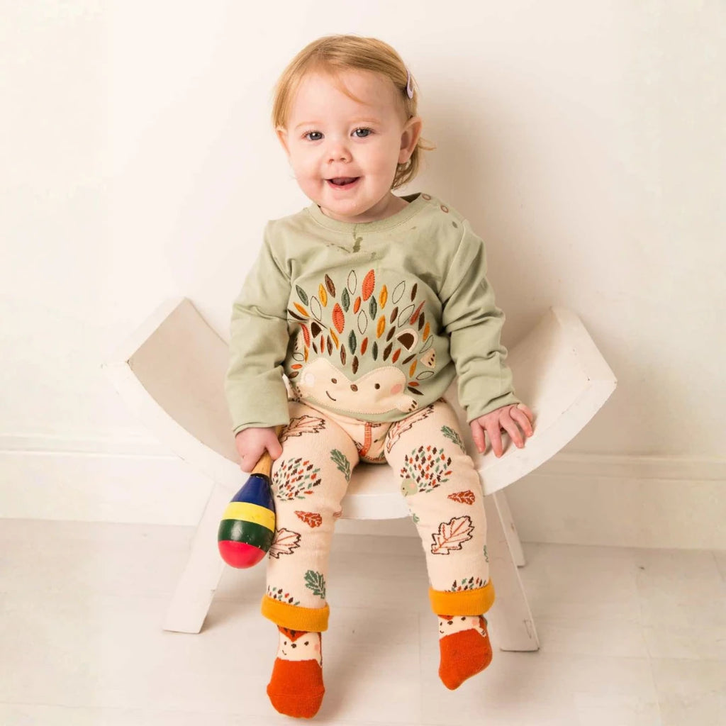 A smiling toddler sits on a small white chair wearing a sage green long-sleeve top with a hedgehog design and beige leggings with colorful leaf prints and orange cuffs. The child holds a small maraca toy against a clean white background.