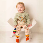 A smiling toddler sits on a small white chair wearing a sage green long-sleeve top with a hedgehog design and beige leggings with colorful leaf prints and orange cuffs. The child holds a small maraca toy against a clean white background.