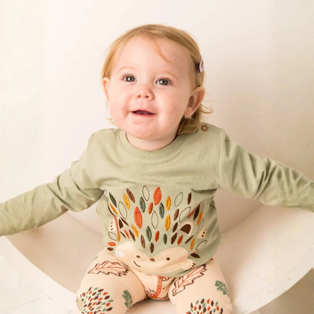 A baby girl wearing a sage green long-sleeve top with a colorful hedgehog design and matching patterned leggings, sitting in a white chair against a warm off-white background.