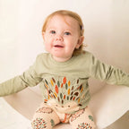 A baby girl wearing a sage green long-sleeve top with a colorful hedgehog design and matching patterned leggings, sitting in a white chair against a warm off-white background.