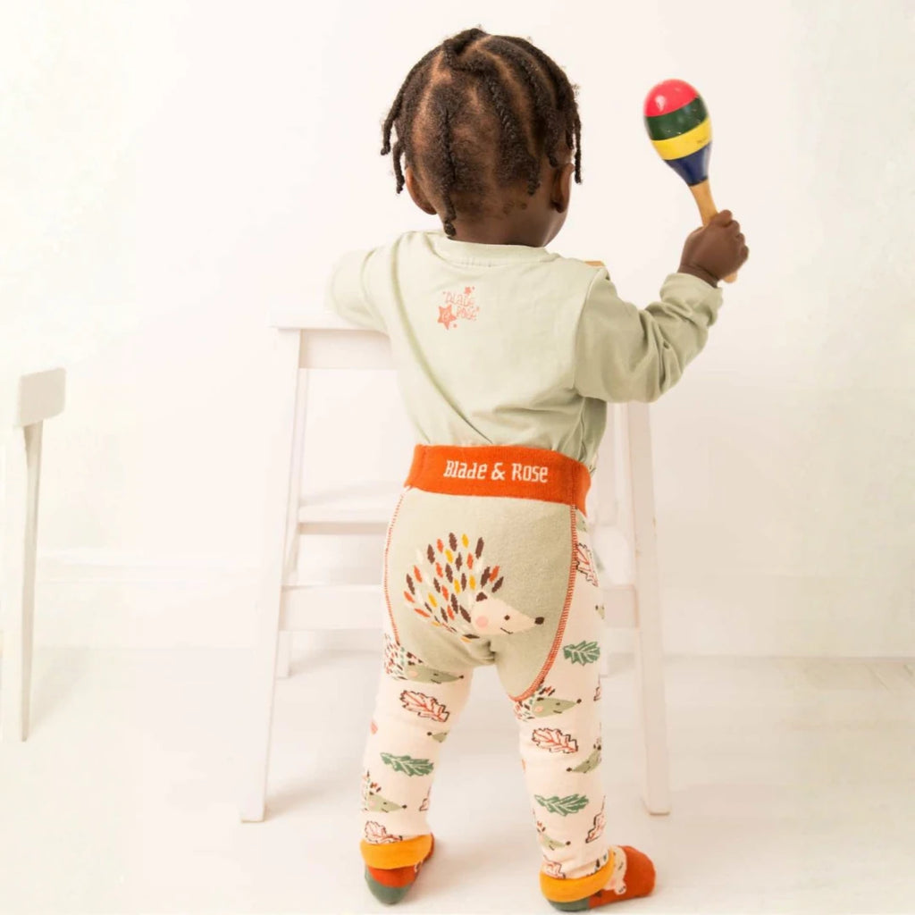 A toddler stands with their back to the camera holding a maraca, wearing a sage green long-sleeve top and patterned leggings with an orange waistband that reads 'Blade & Rose'.