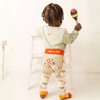 A toddler stands with their back to the camera holding a maraca, wearing a sage green long-sleeve top and patterned leggings with an orange waistband that reads 'Blade & Rose'.