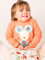 Smiling toddler wearing orange shirt with mouse applique and floral patterned pants sitting on white fur