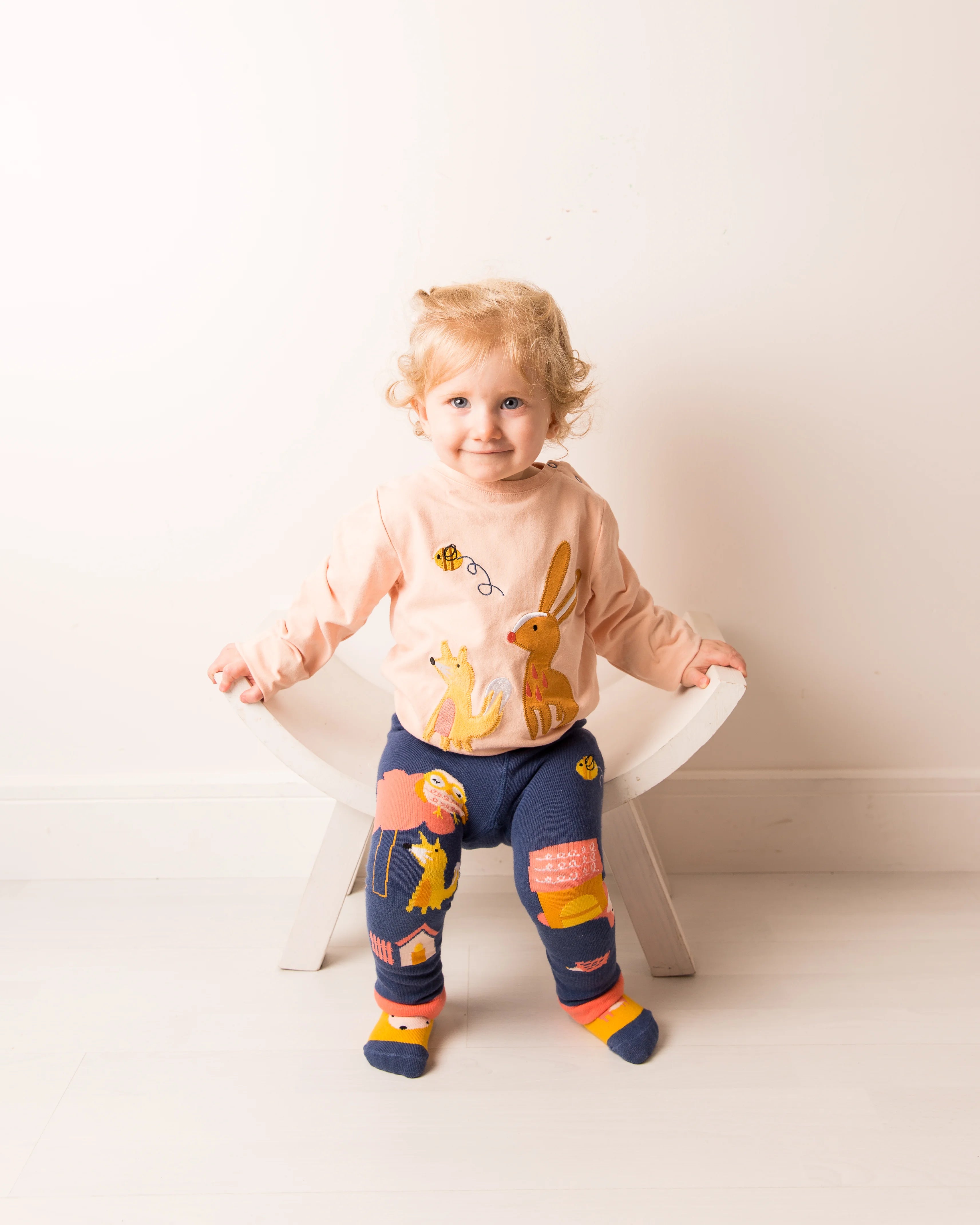 Child wearing a light pink shirt and dark blue pants with colorful animal prints, standing on a white chair.
