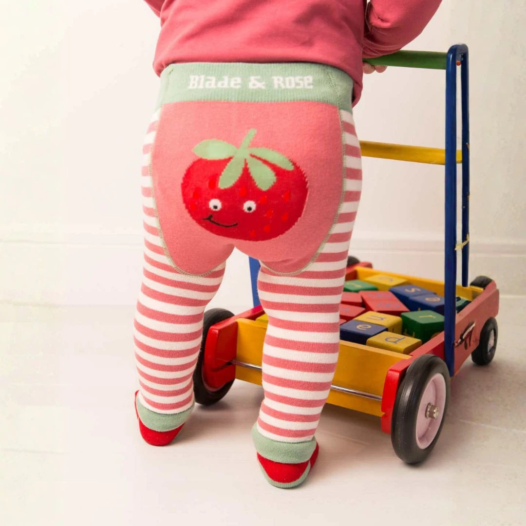 A child wearing pink and white striped tights with a large red strawberry applique on the back and a pink top, standing behind a colorful toy cart.
