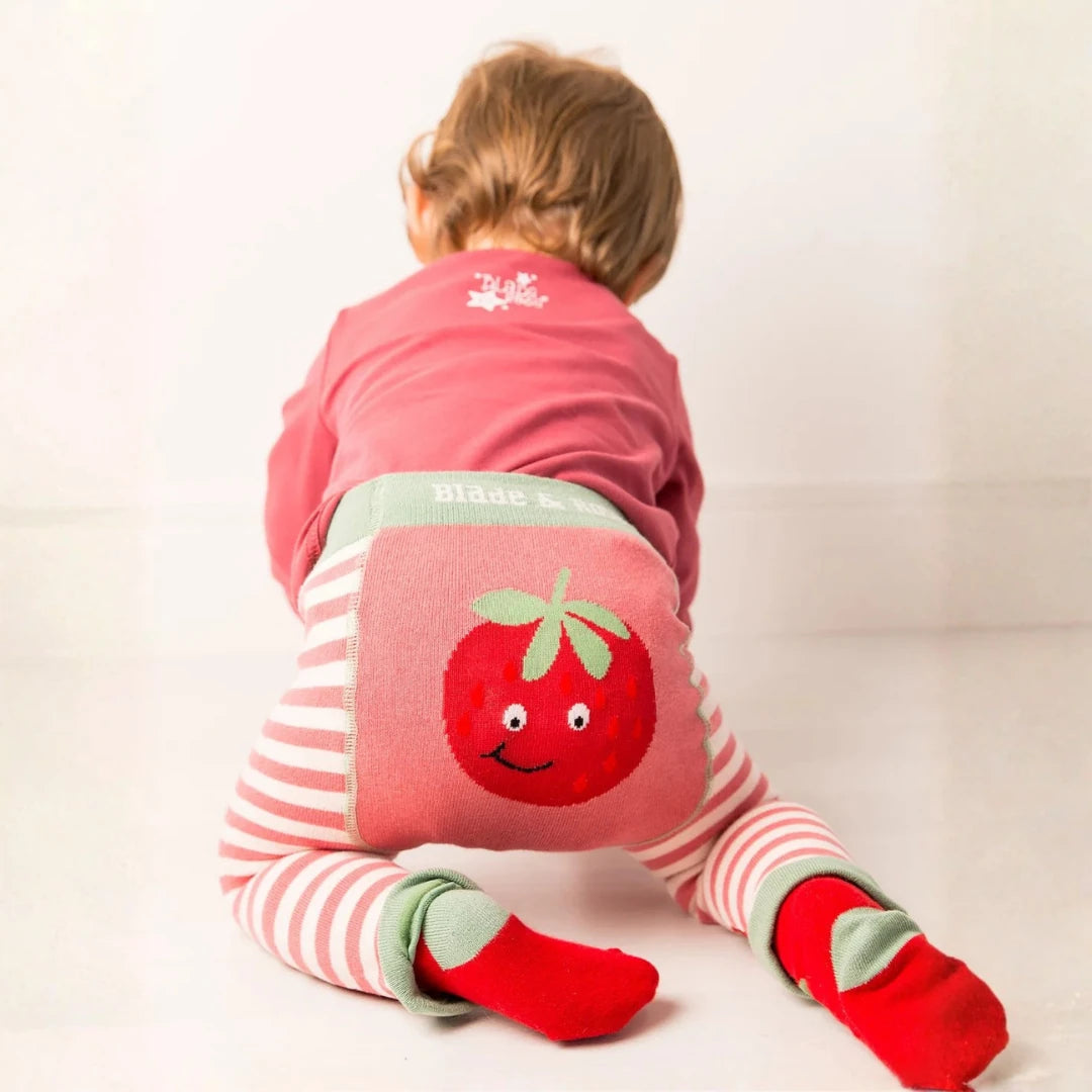 A baby crawling on a white floor, viewed from behind, wearing pink and white striped leggings with a large red strawberry graphic on the back pocket, a pink top, and red socks.