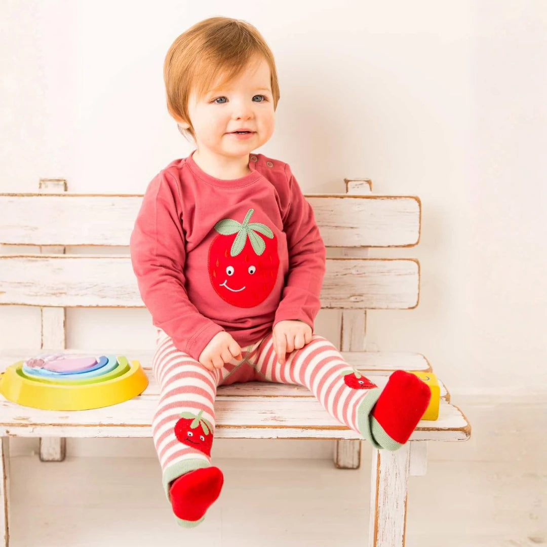 A baby sitting on a rustic white wooden bench wearing a dusty rose long-sleeve top with a smiling red strawberry graphic, matching red and white striped tights with strawberry details, and red socks with green cuffs.