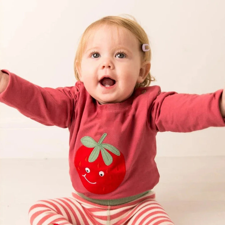 A baby sitting on a light floor with arms outstretched, wearing a pink long-sleeve top with a red strawberry graphic and pink-and-white striped pants with red socks.