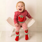 A baby wearing a pink long-sleeve top with a red strawberry graphic and red-and-white striped leggings with matching strawberry patches, sitting on a white wooden chair against a plain white wall.