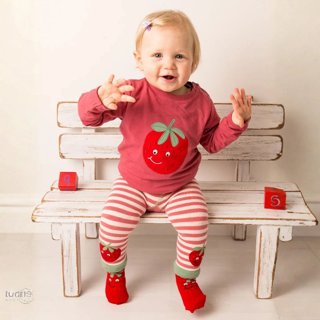A baby girl sitting on a white wooden bench wearing a pink long-sleeve shirt with a strawberry graphic, pink and white striped leggings, and red strawberry socks.