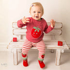 A baby girl sitting on a white wooden bench wearing a pink long-sleeve shirt with a strawberry graphic, pink and white striped leggings, and red strawberry socks.
