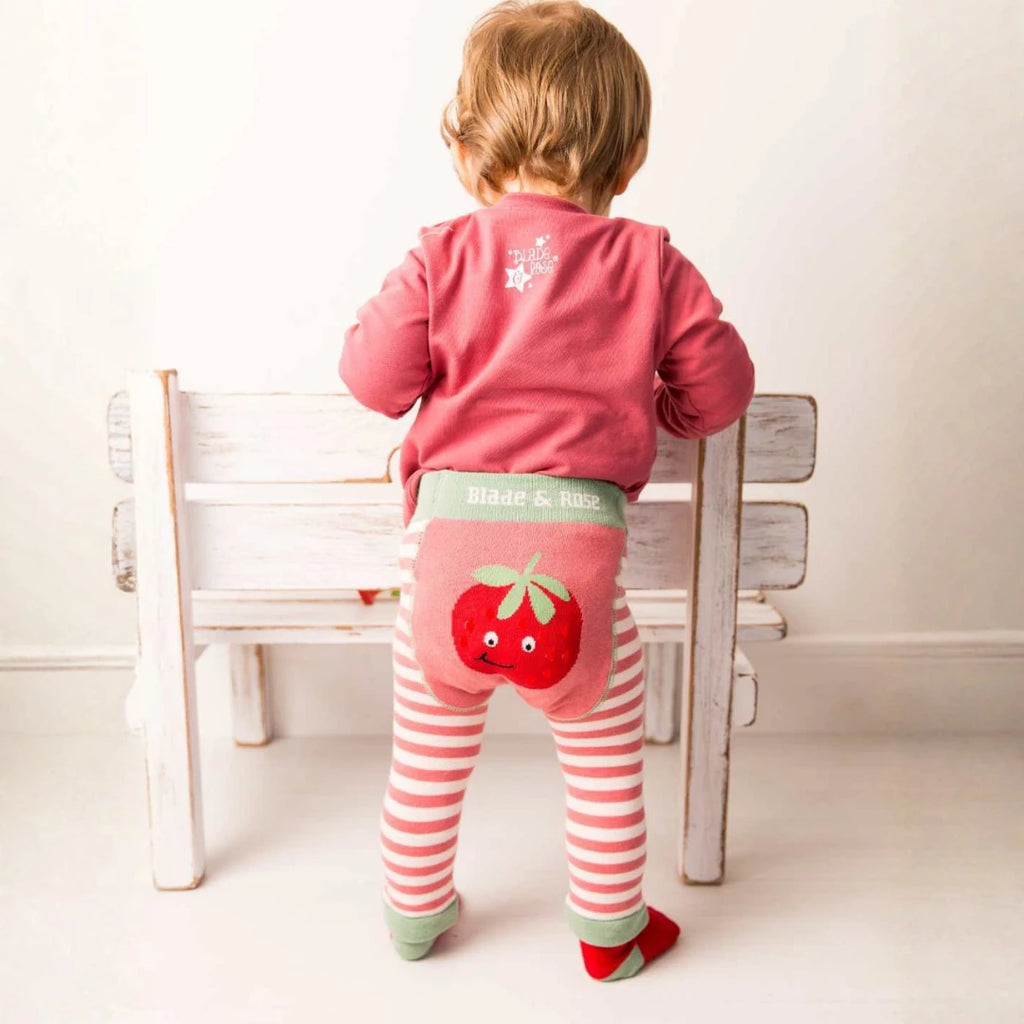 A toddler stands with their back to the camera, leaning on a white wooden bench. The child is wearing a dusty pink long-sleeve top, pink and white striped tights, and red socks.
