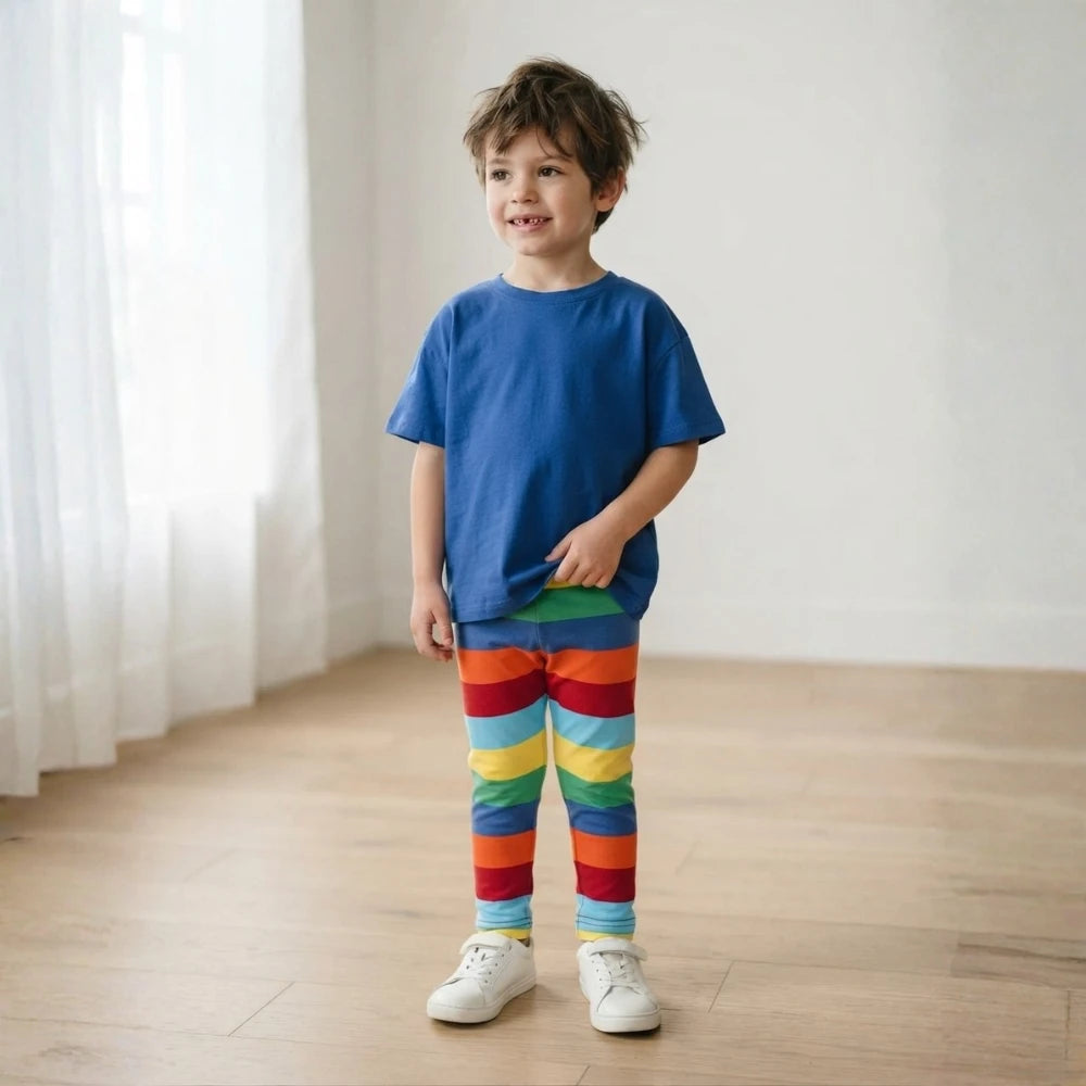 A young boy stands in a bright room wearing a solid blue t-shirt and rainbow-striped leggings with white sneakers.