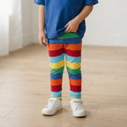 A child wearing rainbow-striped leggings, a blue t-shirt, and white sneakers standing on a light wood floor.