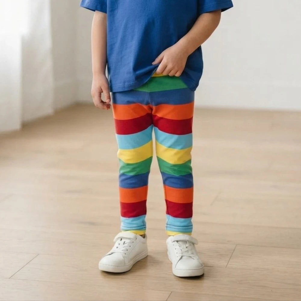 A child wearing rainbow-striped leggings, a blue t-shirt, and white sneakers standing on a light wood floor.