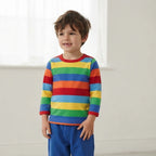 A young boy wearing a long-sleeve rainbow striped shirt and blue pants, standing in a bright, minimalist room with a white window background.