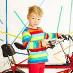 A young boy with blonde hair stands next to a red Raleigh bicycle, wearing a long-sleeve rainbow-striped shirt and red pants, against a white wall with colorful painted lines.