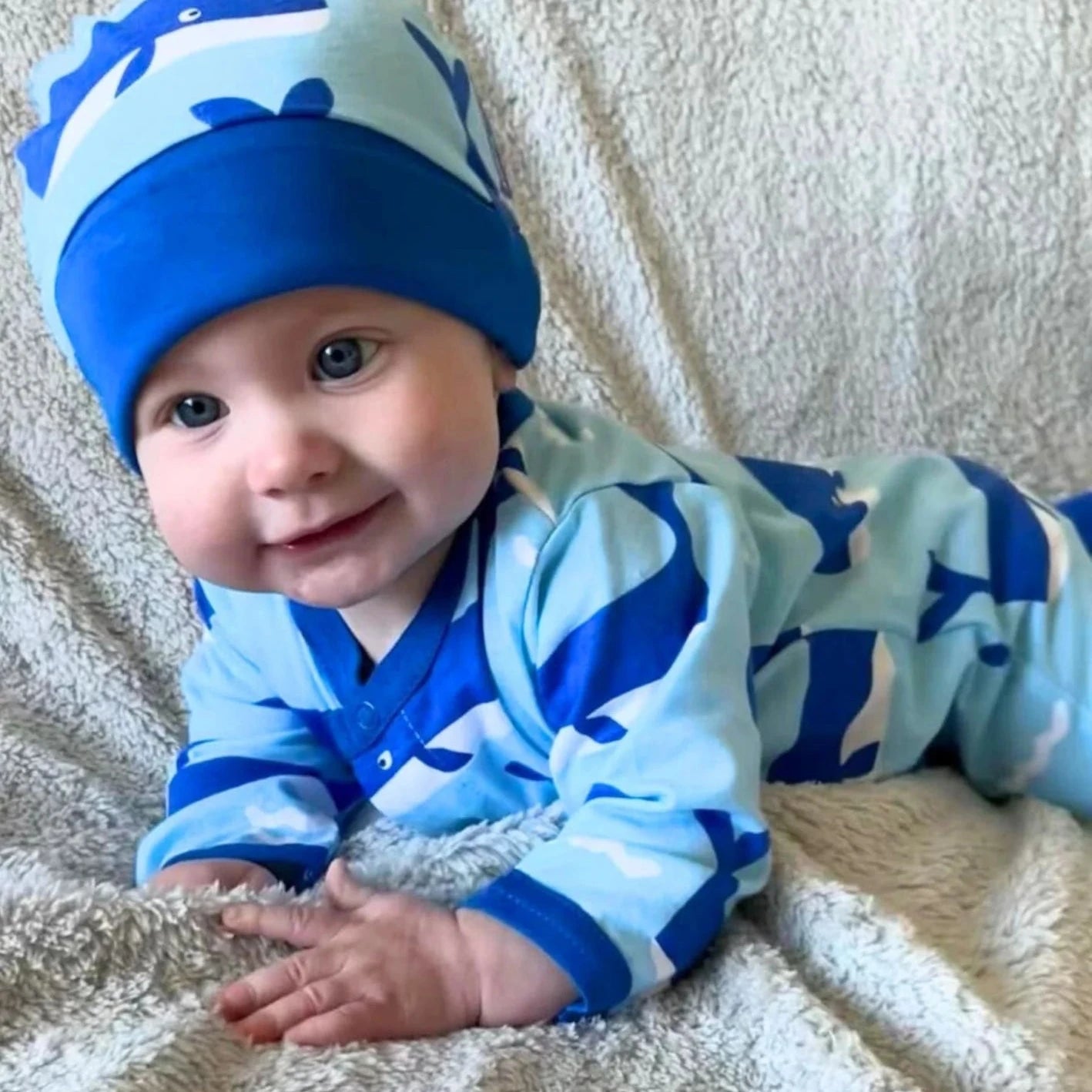 Baby wearing a blue dinosaur-themed outfit and hat on a beige blanket