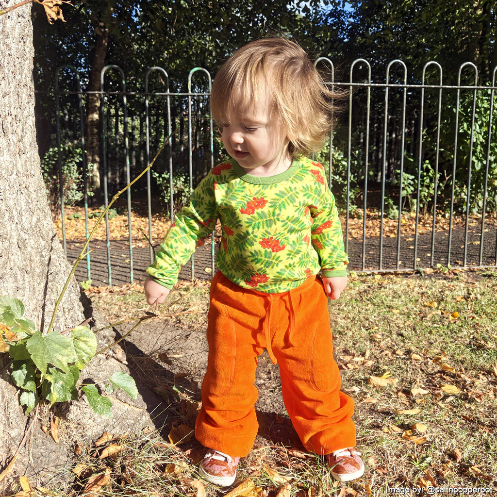 Child wearing a green floral shirt and orange pants standing outdoors near a tree and fence.