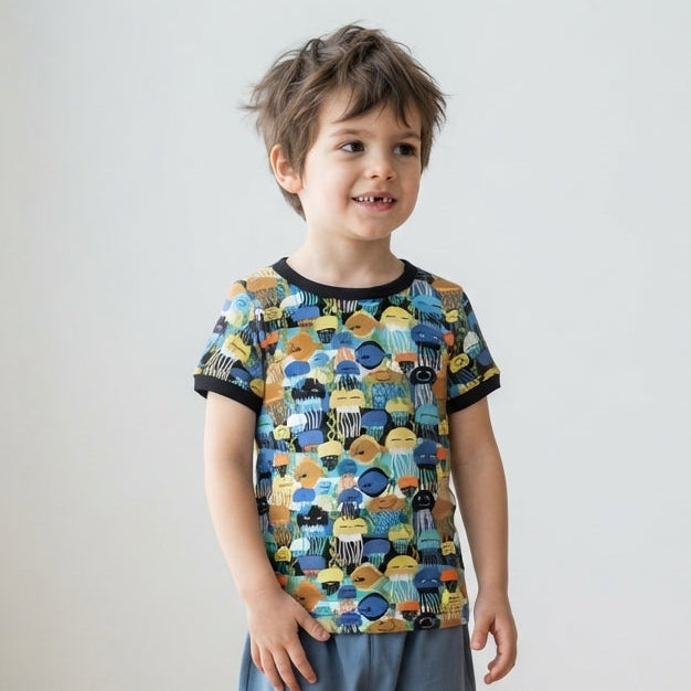 A young child standing in a bright room with light wood floors and white walls, wearing a short-sleeve ringer T-shirt with a colorful fish pattern and blue pants.