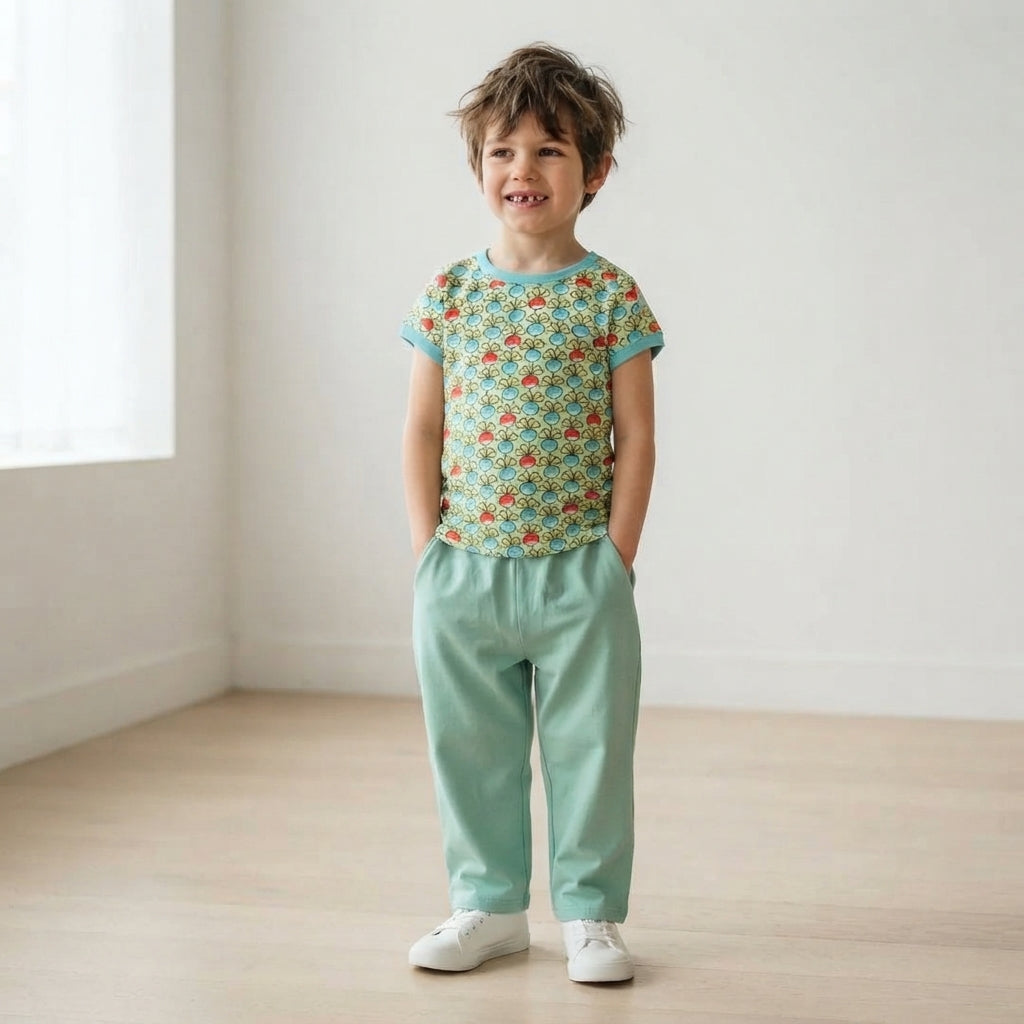 A young boy stands in a bright, minimalist room wearing a short-sleeve t-shirt with a colorful geometric pattern and mint green pants.