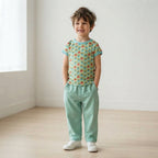 A young boy stands in a bright, minimalist room wearing a short-sleeve t-shirt with a colorful geometric pattern and mint green pants.