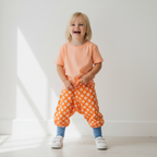 Child wearing an orange shirt and patterned pants standing against a white background