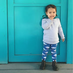 Child wearing a gray sweatshirt and blue leggings with white patterns, standing in front of a teal door.